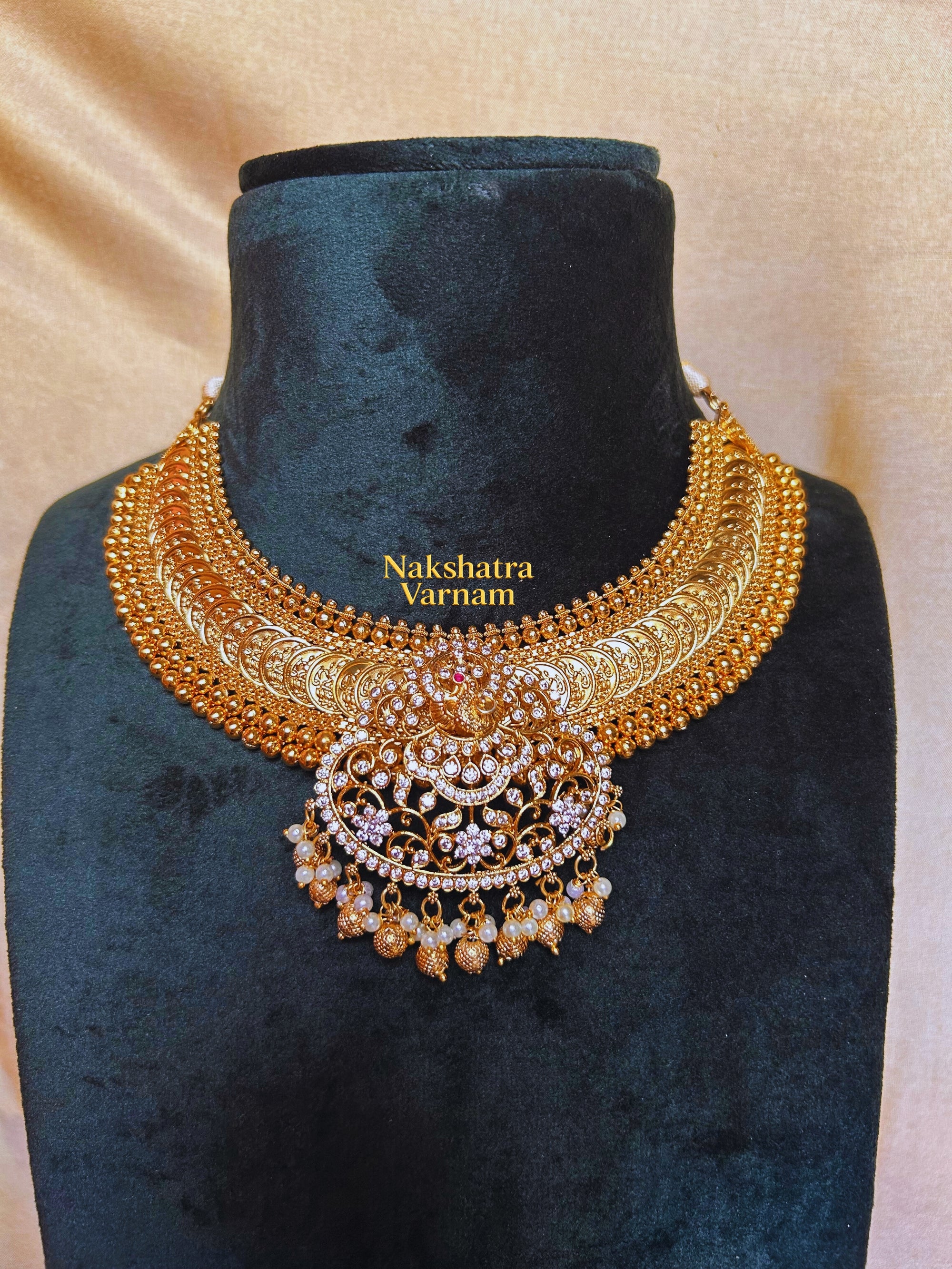 Gold necklace on a black velvet stand with 'Nakshatra Varnam' branding.